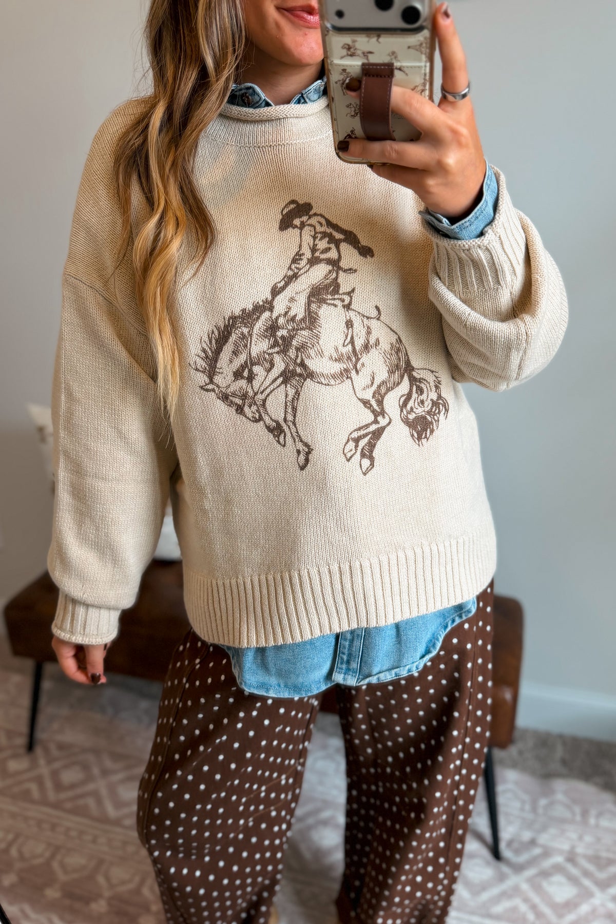 A woman takes a mirror selfie wearing a cream-colored knit sweater with a brown cowboy riding a bucking horse graphic, layered over a blue denim shirt and paired with brown polka-dot pants.