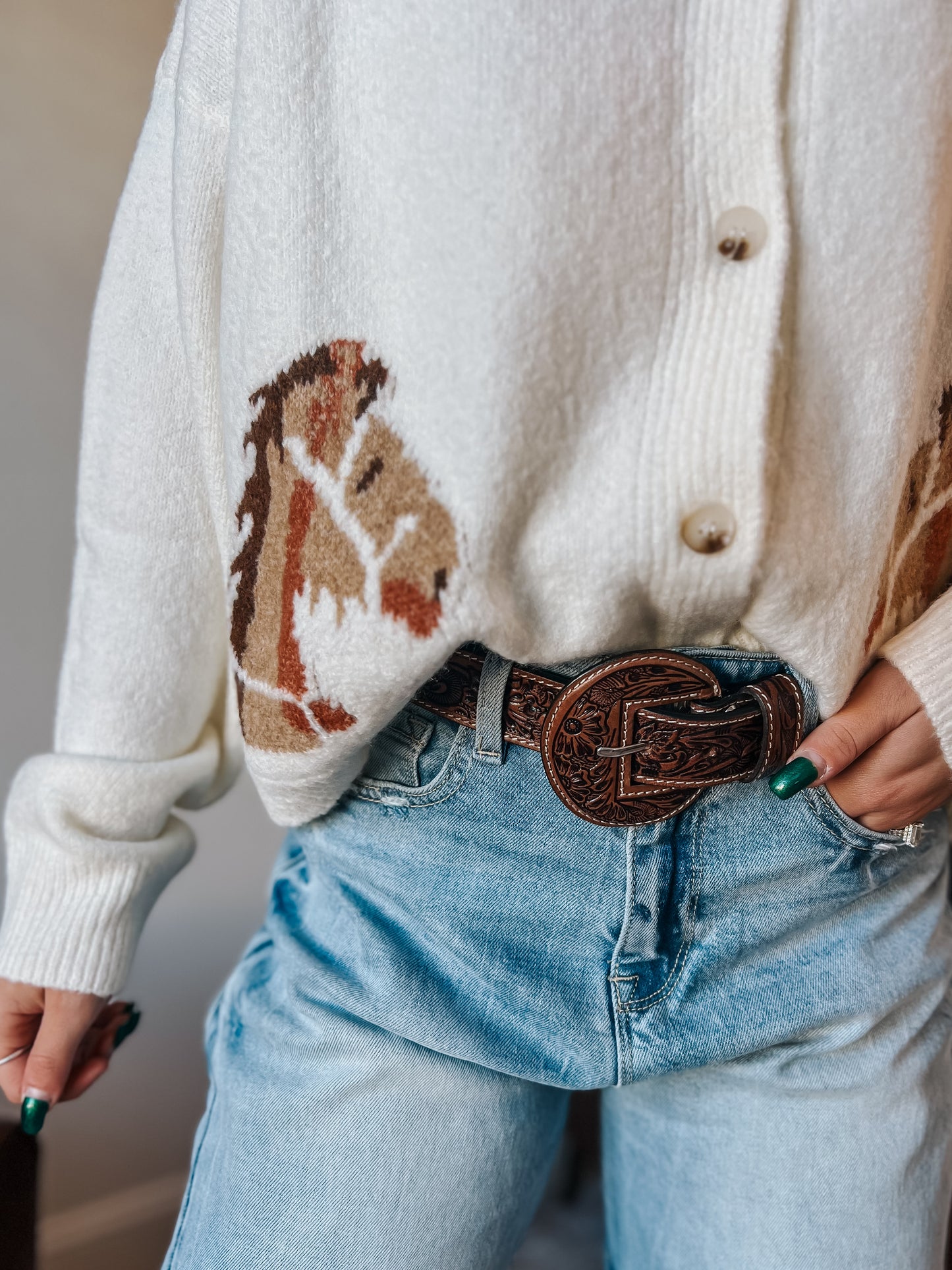Person wearing a white cardigan with a horse design, blue jeans, and a brown belt.