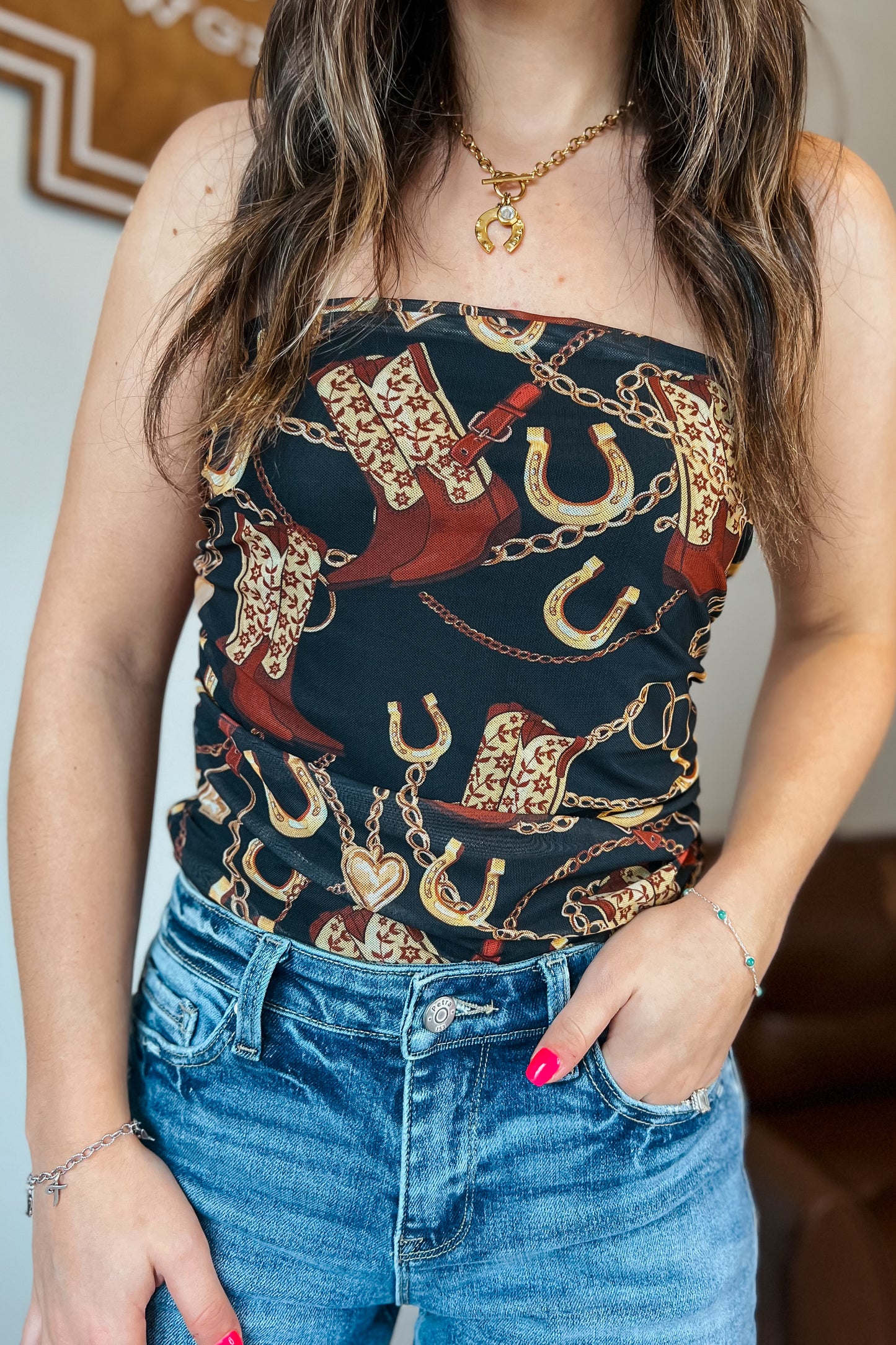 Strapless black western print top featuring cowboy boots, horseshoes, and gold chain details, shown styled with blue jeans.