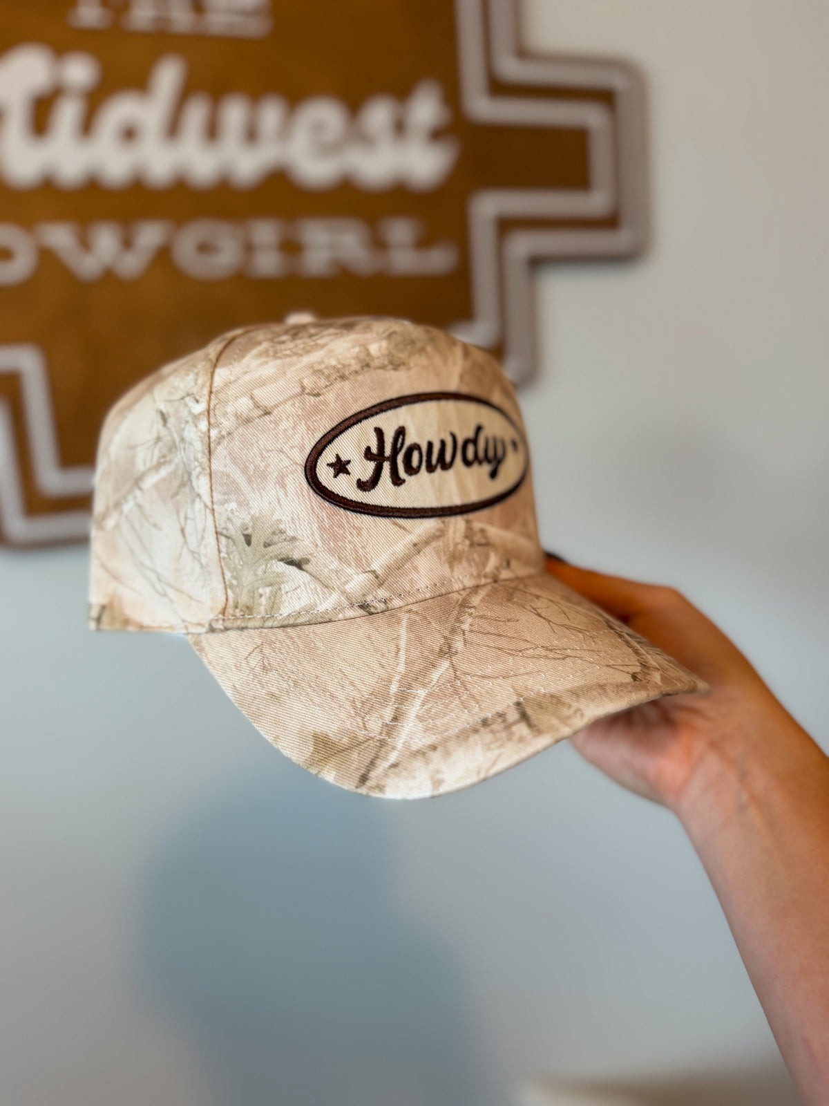 A camouflage baseball cap held by a hand, featuring an oval embroidered patch on the front that reads 'Howdy' with stars on each side. The background shows a light-colored wall and a wooden sign.