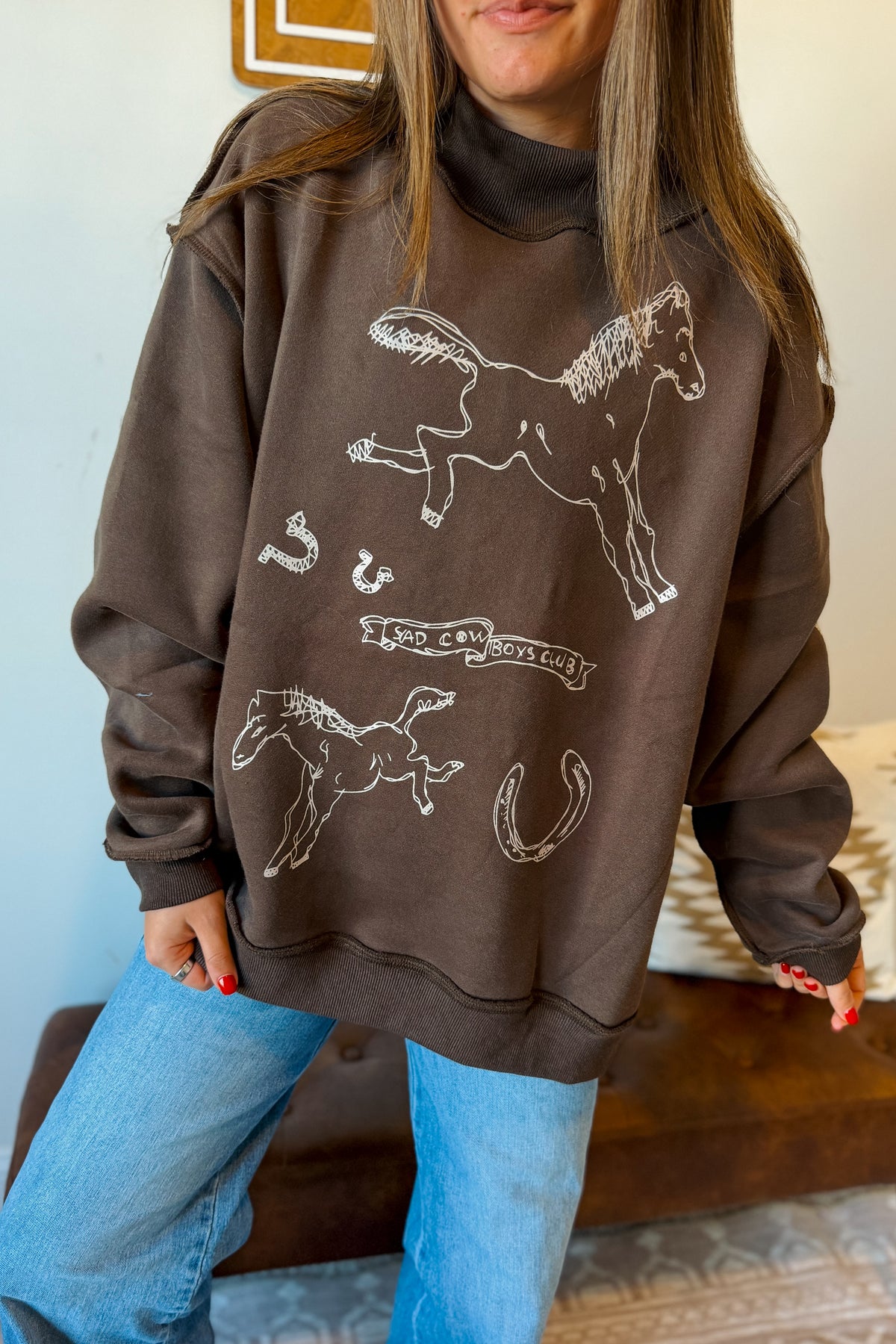 A woman wearing a dark brown sweatshirt with white horse line drawings and blue jeans, standing indoors.