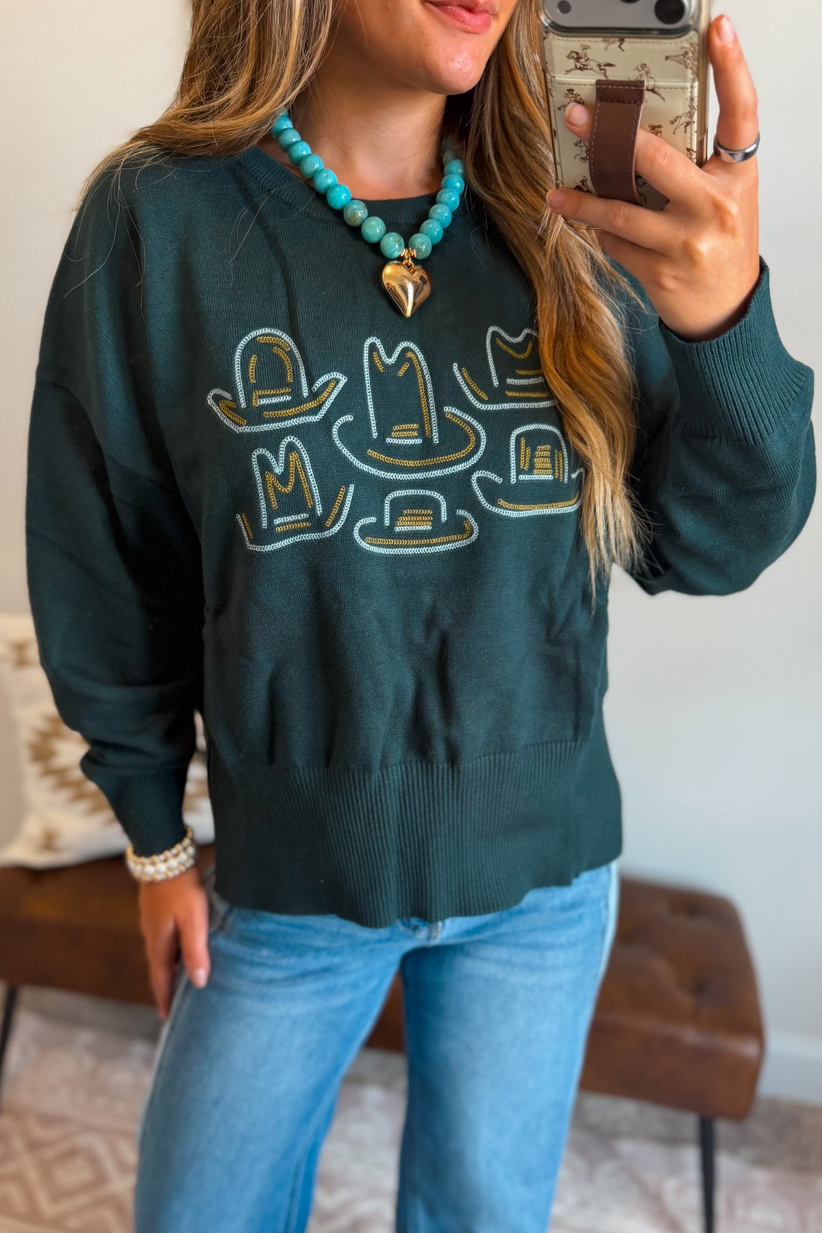 A woman takes a mirror selfie wearing a dark teal crewneck sweatshirt with embroidered cowboy hats, paired with light blue jeans and a turquoise beaded necklace.
