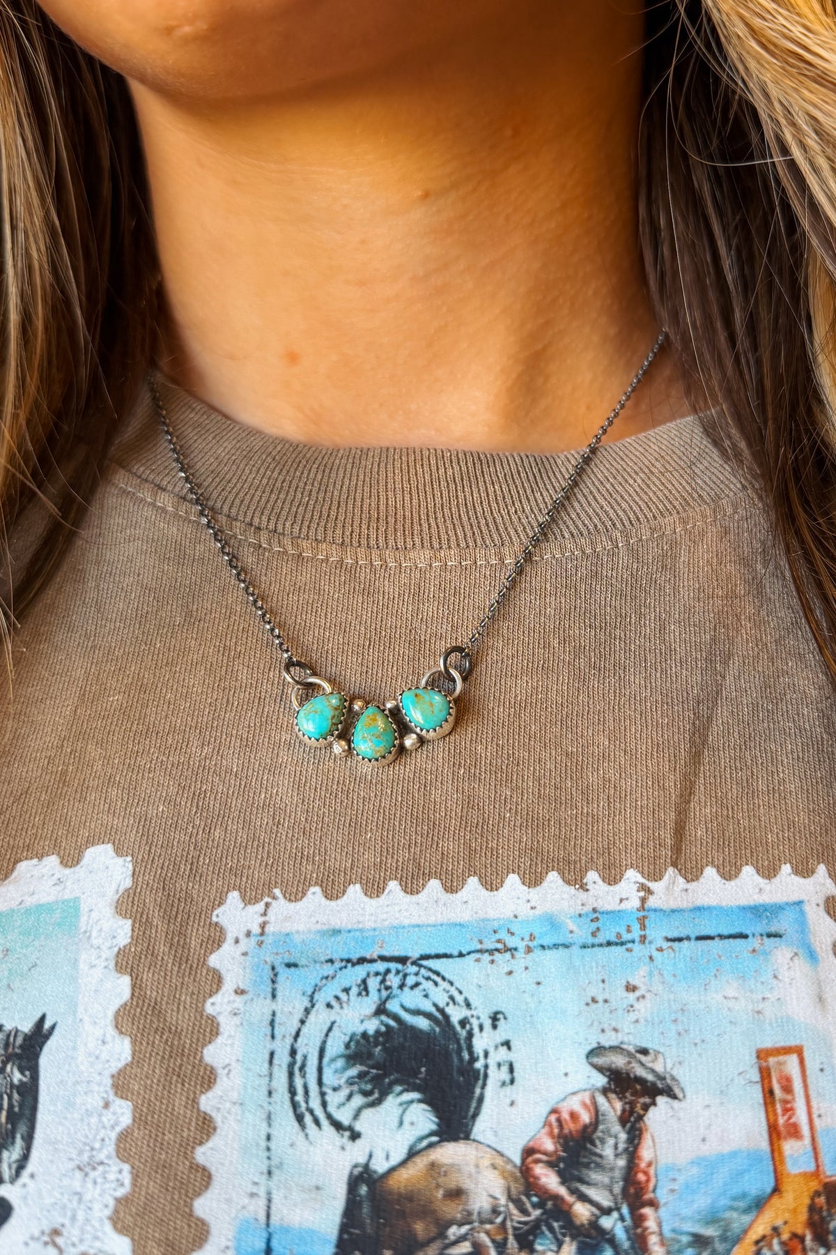 A close-up of a person wearing a silver-toned necklace with three turquoise-colored stones, styled over a brown graphic t-shirt.