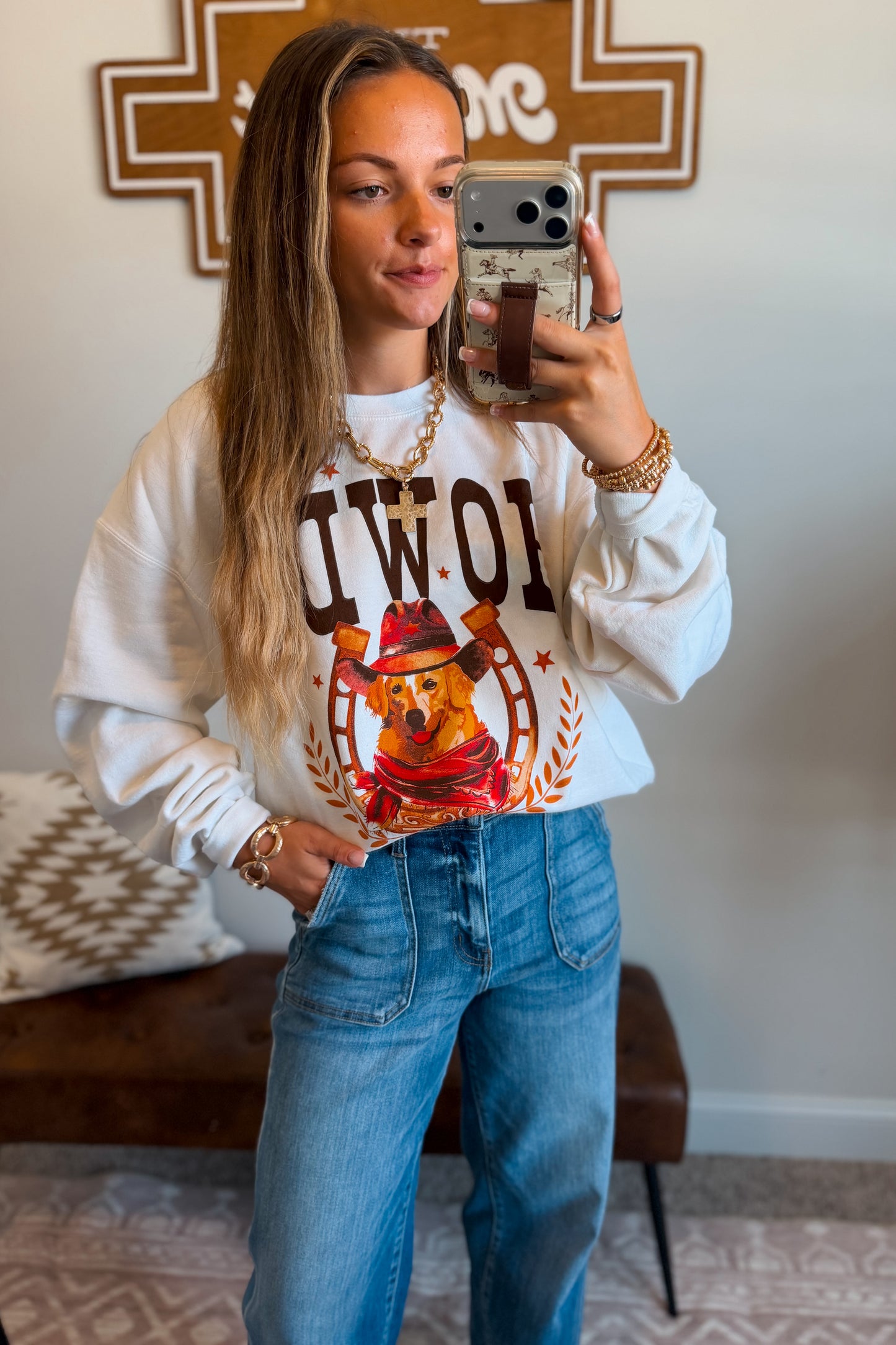 A woman takes a mirror selfie wearing a white crewneck sweatshirt with brown lettering and a cowboy dog graphic, paired with blue jeans.