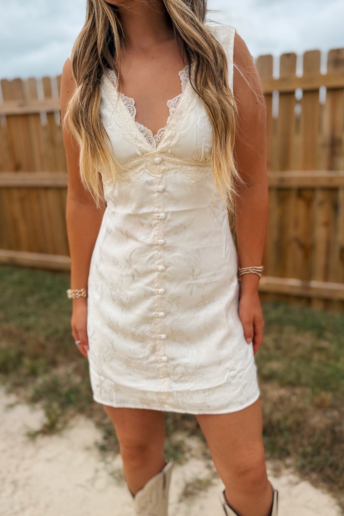 A woman wearing a sleeveless cream-colored lace and textured mini dress with a button-down front, standing outdoors in front of a wooden fence.