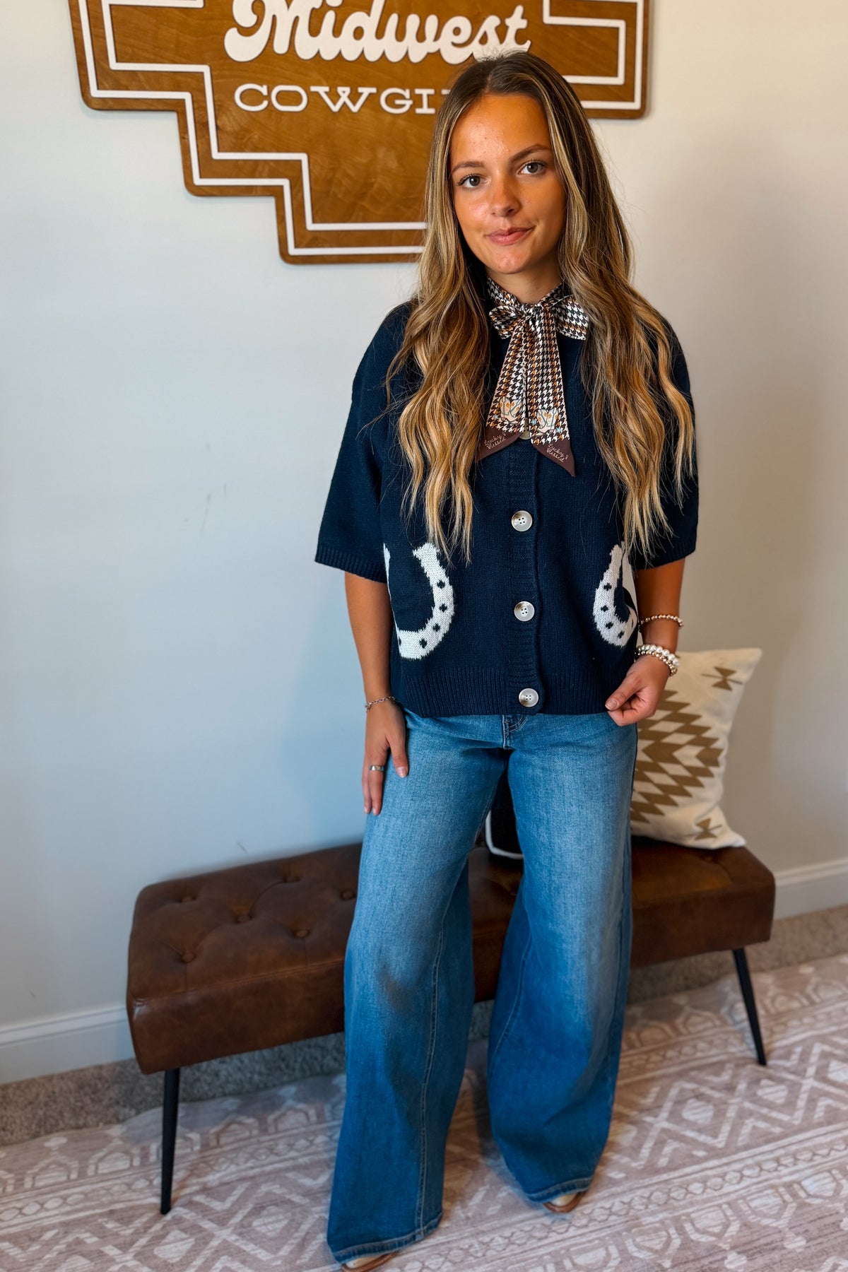 A woman stands indoors wearing a navy short-sleeve button-front top with white horseshoe motifs on the pockets, a plaid neck scarf, and wide-leg blue jeans.