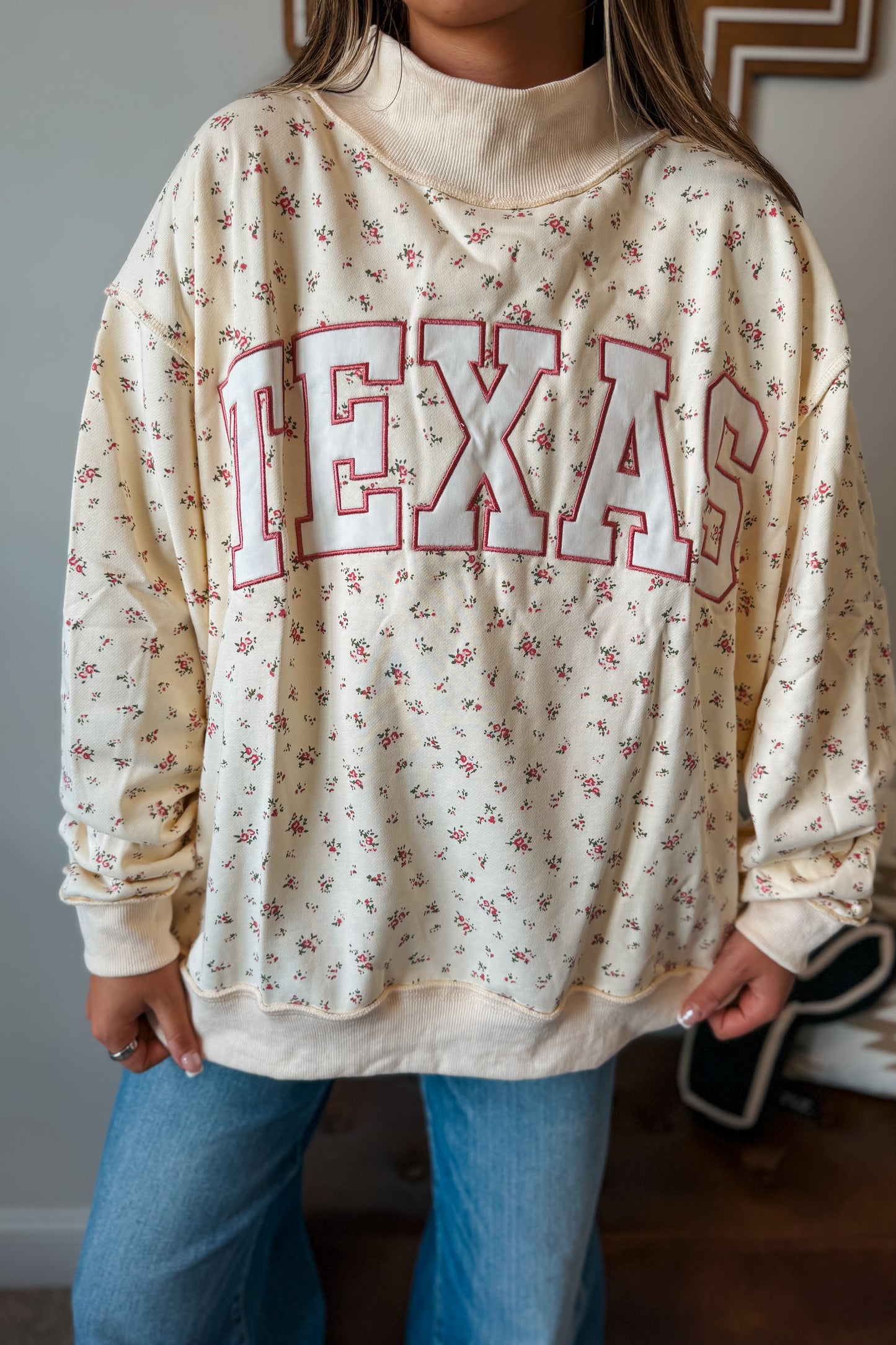 A person wearing a cream-colored long-sleeve sweatshirt with a small pink floral print and 'TEXAS' written across the front in large outlined letters.
