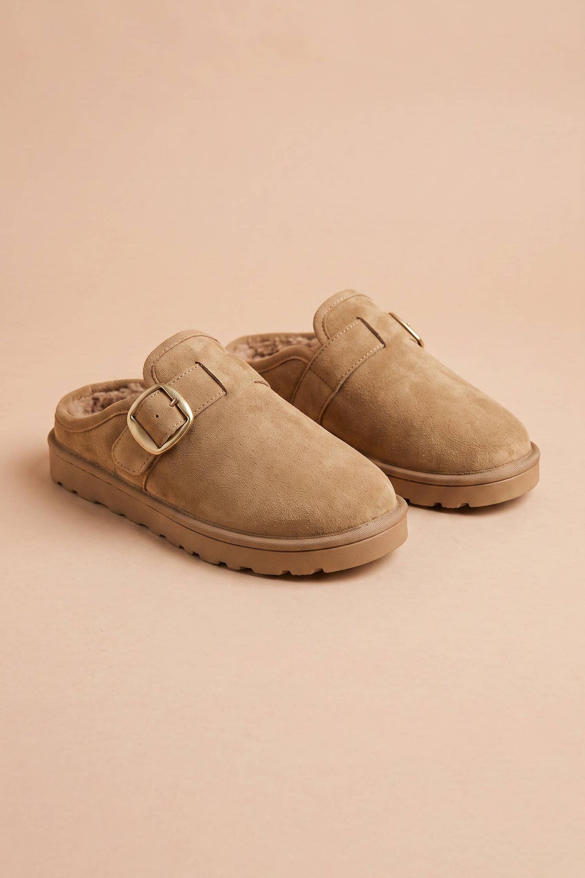 A pair of beige suede clogs with adjustable buckle straps, displayed on a solid beige background.