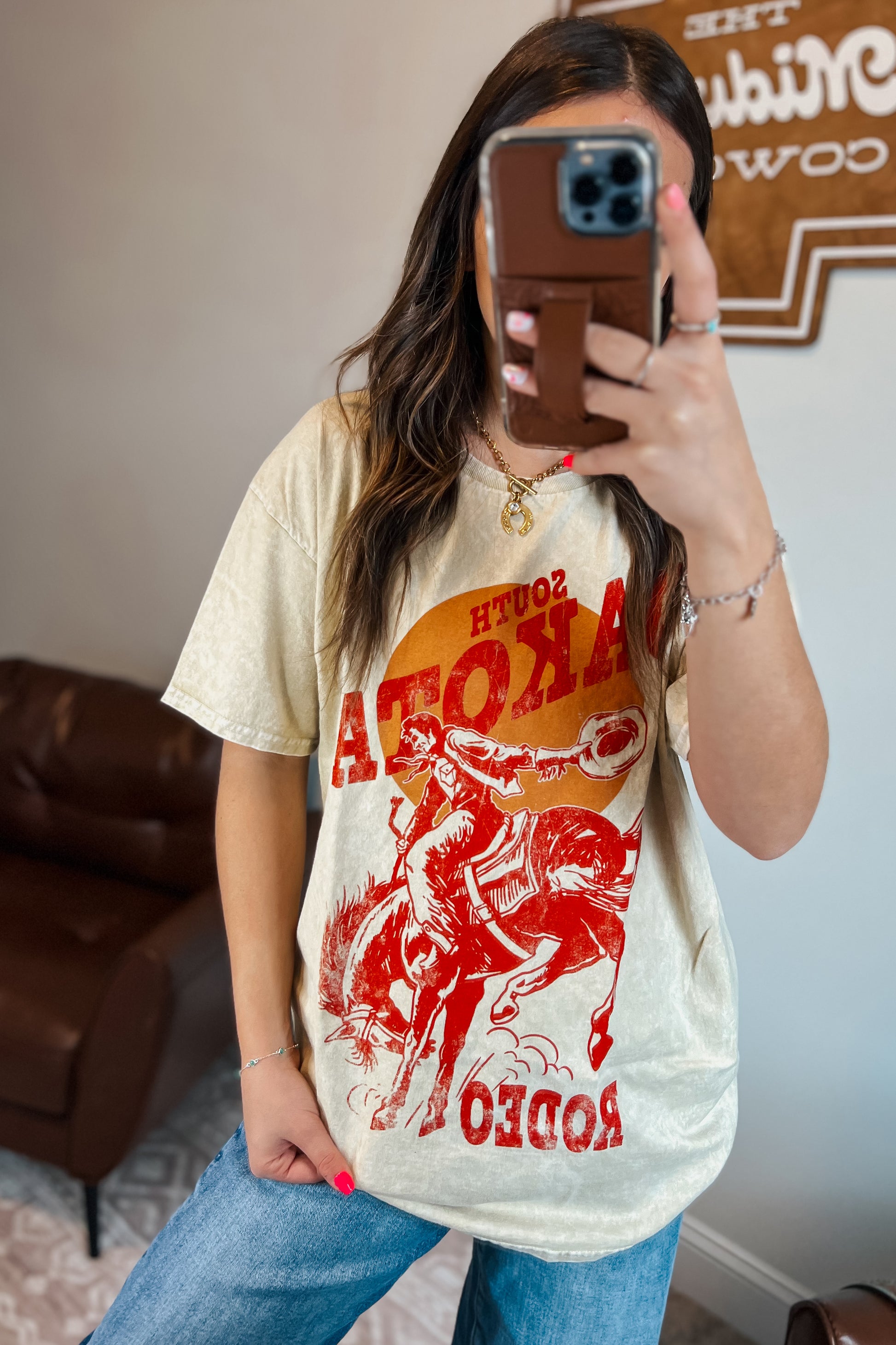 Light tan mineral-wash T-shirt with red “South Dakota Rodeo” graphic featuring a cowboy riding a bucking horse inside an orange sunset design