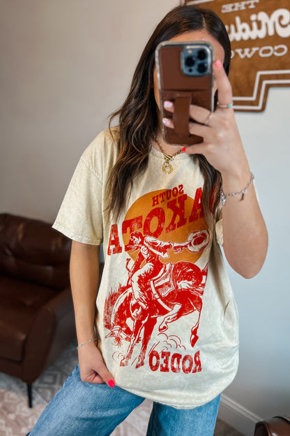 Light tan mineral-wash T-shirt with red “South Dakota Rodeo” graphic featuring a cowboy riding a bucking horse inside an orange sunset design
