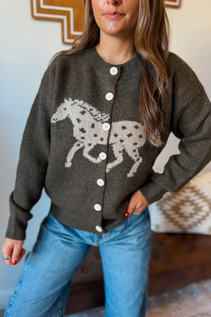 A woman wearing a dark olive brown knit cardigan sweater with a large beige and black spotted horse graphic on the front, paired with blue jeans.
