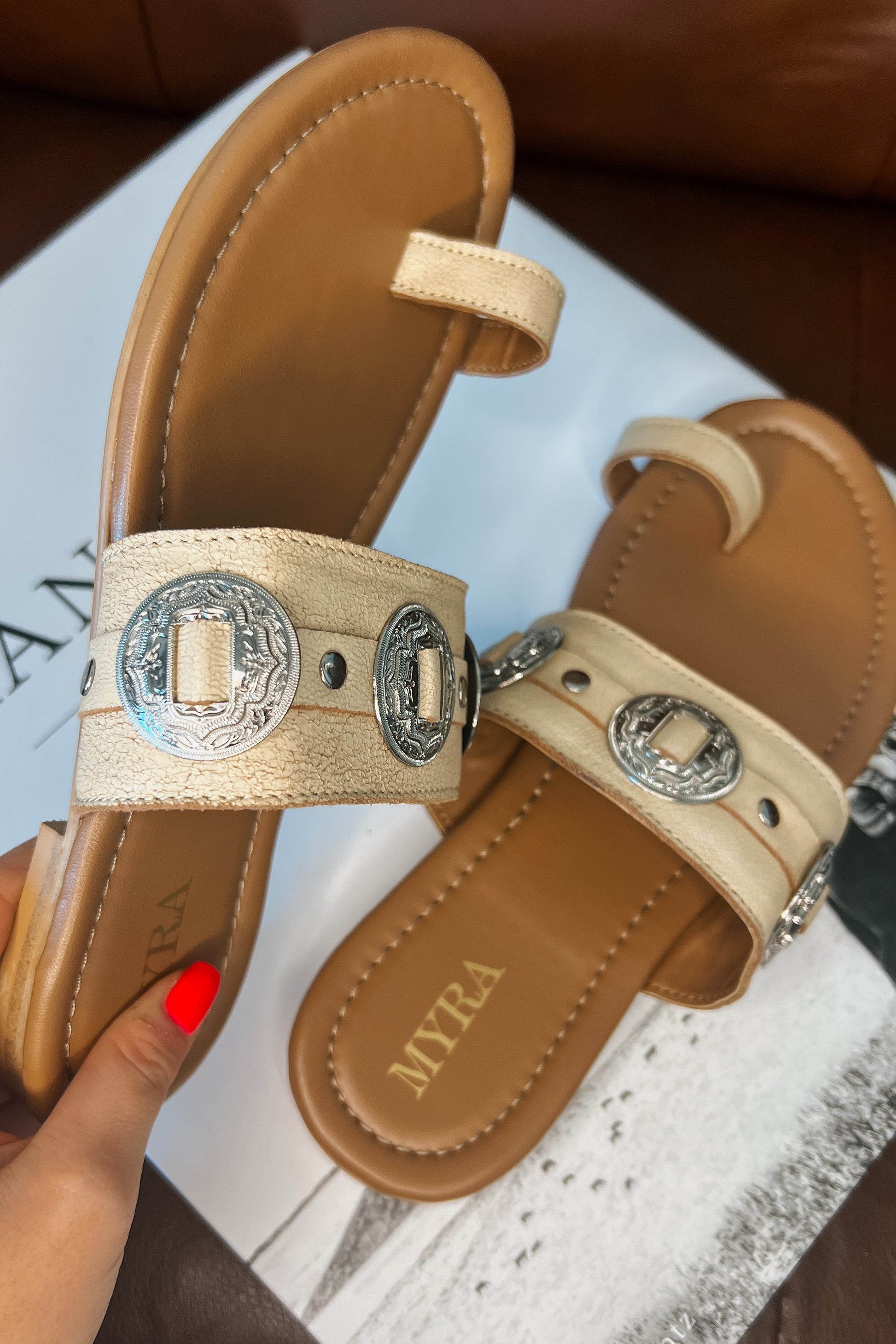 Cream western slide sandals with tan cushioned footbed and toe loop. Wide textured strap features silver engraved concho accents and metal studs. Brand name printed on footbed.
