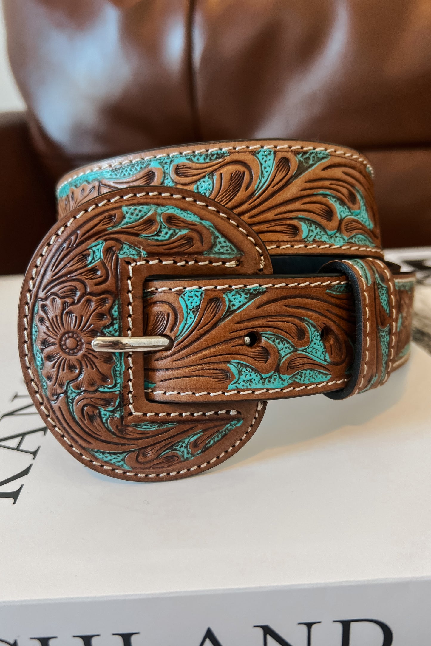 Tan leather western belt with turquoise inlay and floral tooling, featuring a round engraved buckle, styled with blue jeans and a brown top.