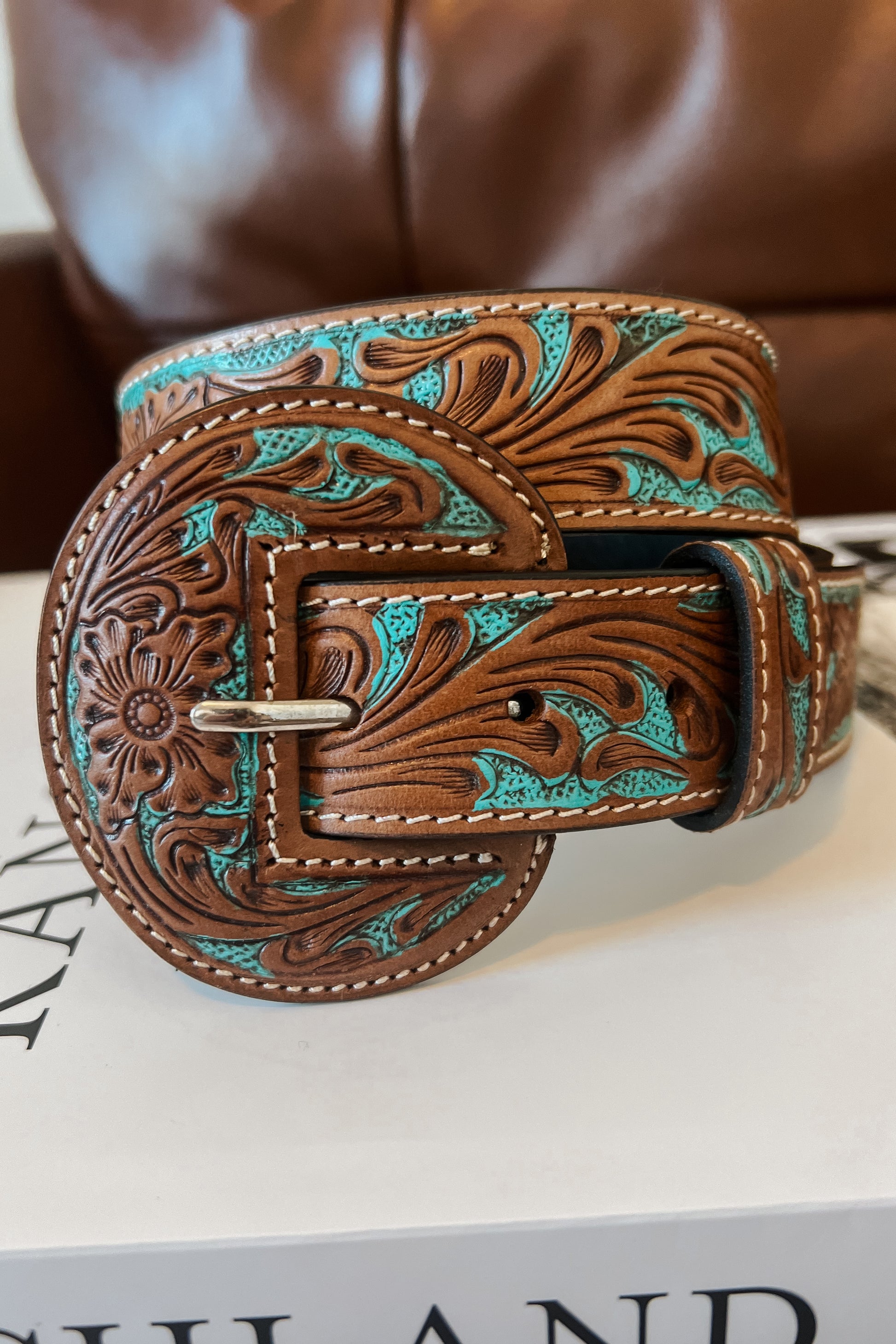 Tan leather western belt with turquoise inlay and floral tooling, featuring a round engraved buckle, styled with blue jeans and a brown top.