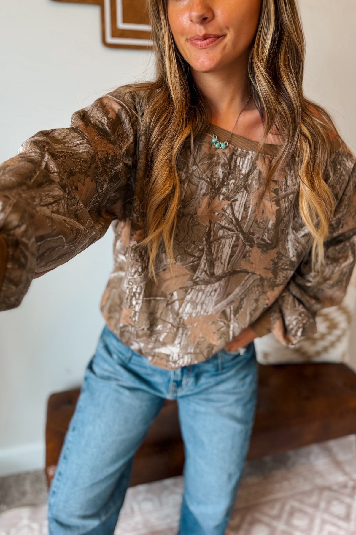 A front selfie of woman wearing a brown and beige camouflage-patterned off-the-shoulder sweatshirt, blue jeans, 