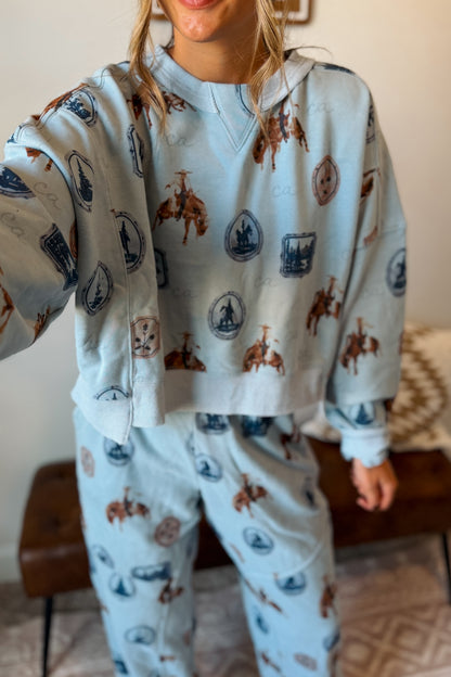 A person wearing a light blue matching lounge set with an allover western-themed print featuring brown cowboy boots and blue horseshoes, photographed indoors.