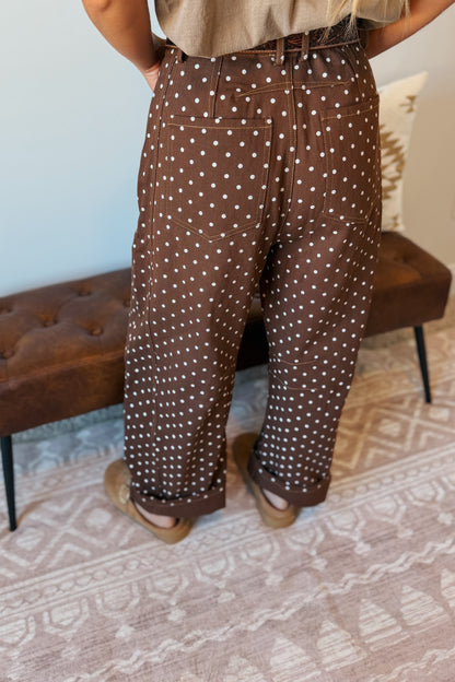 A person shown from the waist down wearing brown wide-leg pants with a white polka dot pattern, standing on a patterned rug in front of a tufted bench.