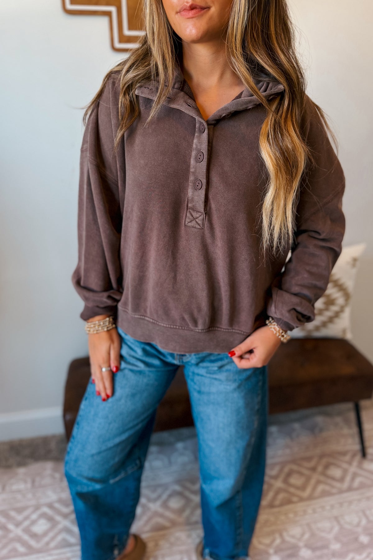 A woman wearing a brown long-sleeve button-up henley sweatshirt and blue jeans, standing in a home interior setting.