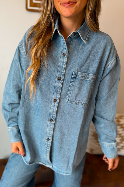A woman wearing an oversized light blue denim button-up shirt with a chest pocket, paired with matching blue jeans in an indoor setting.