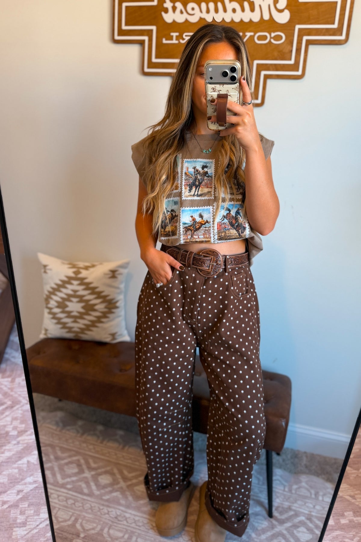 A woman takes a mirror selfie showing an outfit consisting of a brown graphic crop top and brown polka dot wide-leg pants with a matching belt, paired with tan boots. The background shows a home interior with a bench, patterned pillow, and a wooden sign.