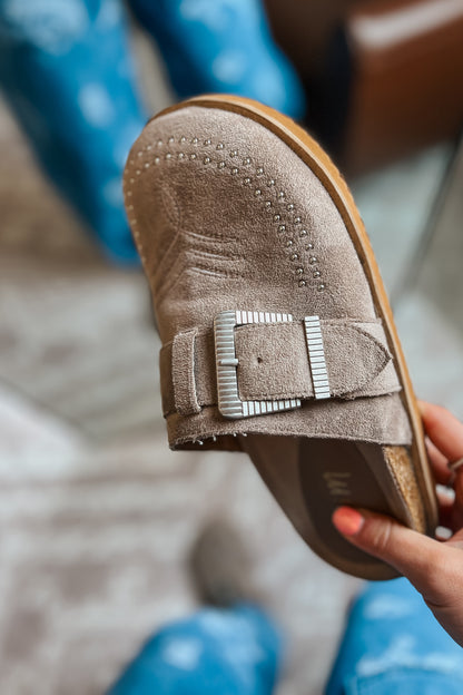 Taupe suede western mule with silver buckle strap, studded toe detail, and low-profile slip-on design.