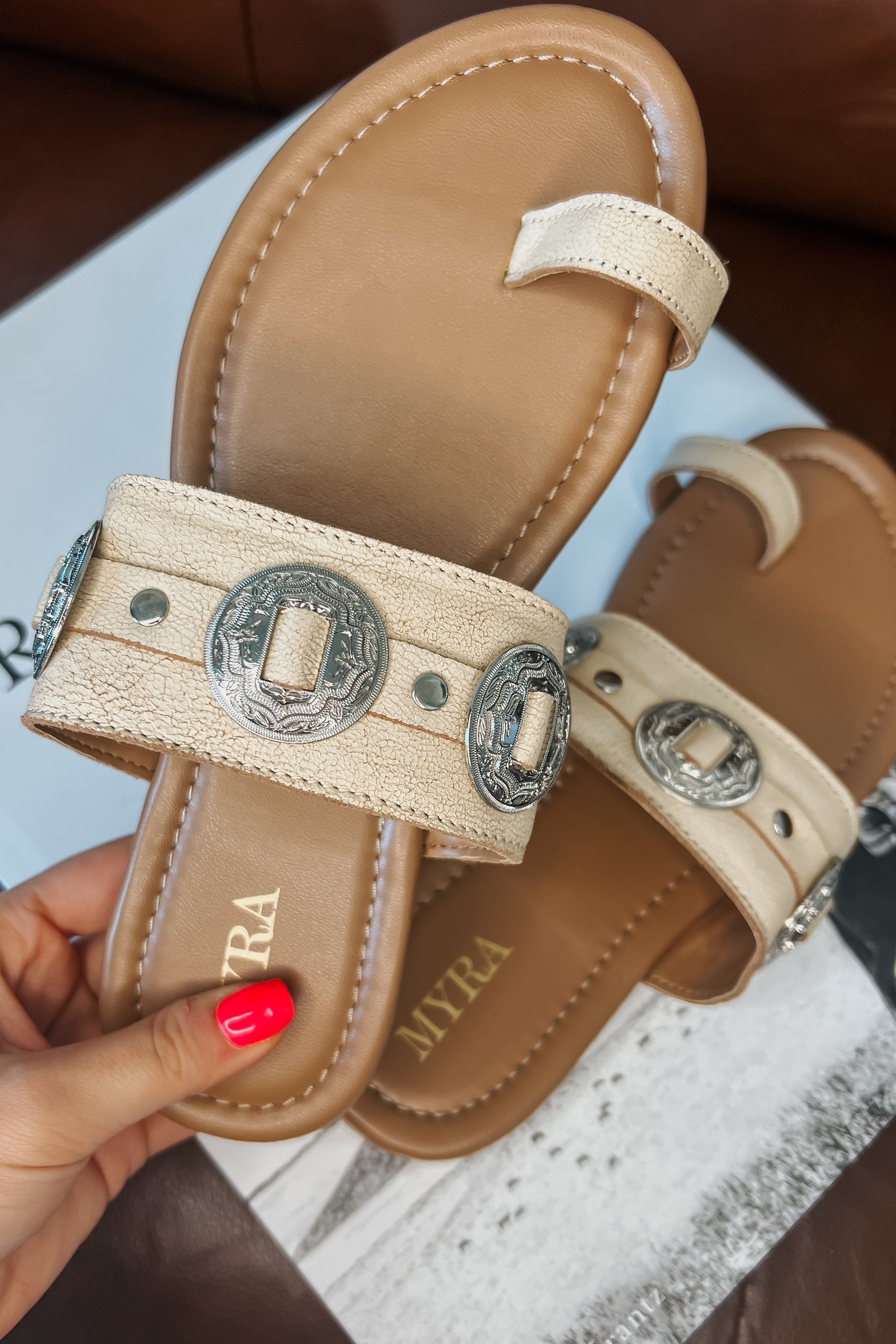 Cream western slide sandals with tan cushioned footbed and toe loop. Wide textured strap features silver engraved concho accents and metal studs. Brand name printed on footbed.