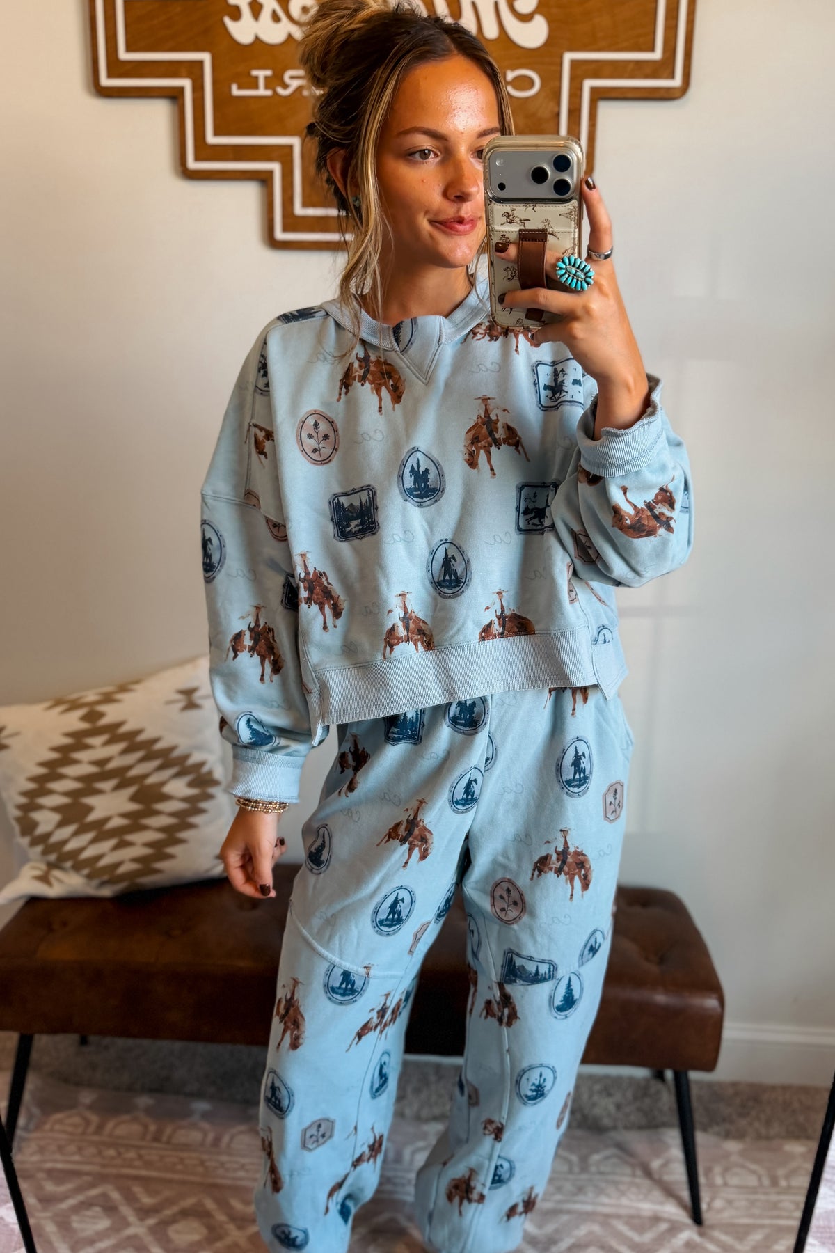 A woman takes a mirror selfie wearing a light blue matching lounge set with a cowboy and Indian head print. The set includes a long-sleeve sweatshirt and sweatpants.