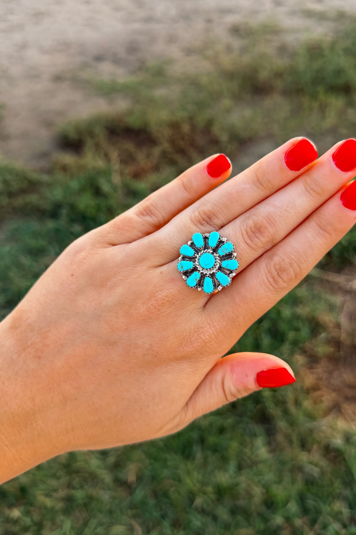 A close-up of a hand with bright red nail polish wearing a silver-toned ring with a turquoise and black floral pattern, set against a blurred outdoor background of grass and sand.