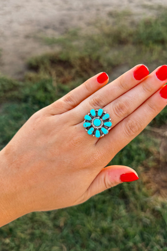 A close-up of a hand with bright red nail polish wearing a silver-toned ring with a turquoise and black floral pattern, set against a blurred outdoor background of grass and sand.