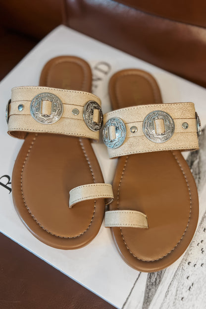 Cream western slide sandals with tan cushioned footbed and toe loop. Wide textured strap features silver engraved concho accents and metal studs. Brand name printed on footbed.