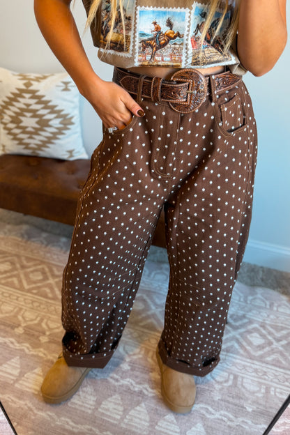 A person wearing brown barrel-leg pants with a white polka dot pattern, styled with a brown leather belt and tan boots in an indoor setting.