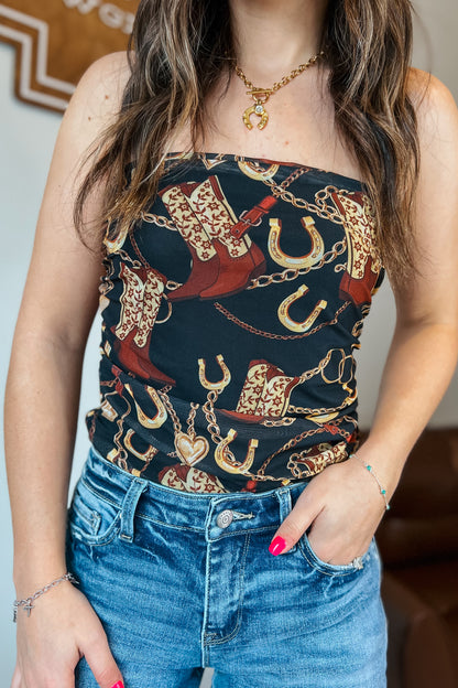 Strapless black western print top featuring cowboy boots, horseshoes, and gold chain details, shown styled with blue jeans.
