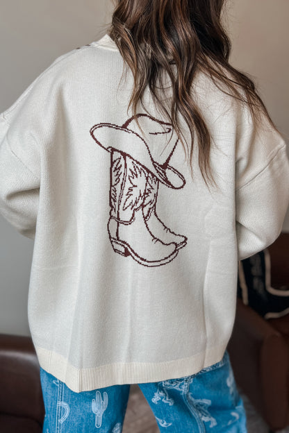 Back view of a cream knit cardigan featuring a brown cowboy boot and hat graphic on the back, styled with blue western print jeans.