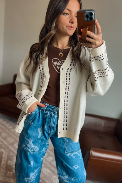 Model wearing a cream knit cardigan with brown western-inspired stitch detailing over a brown graphic tee and blue western print jeans.