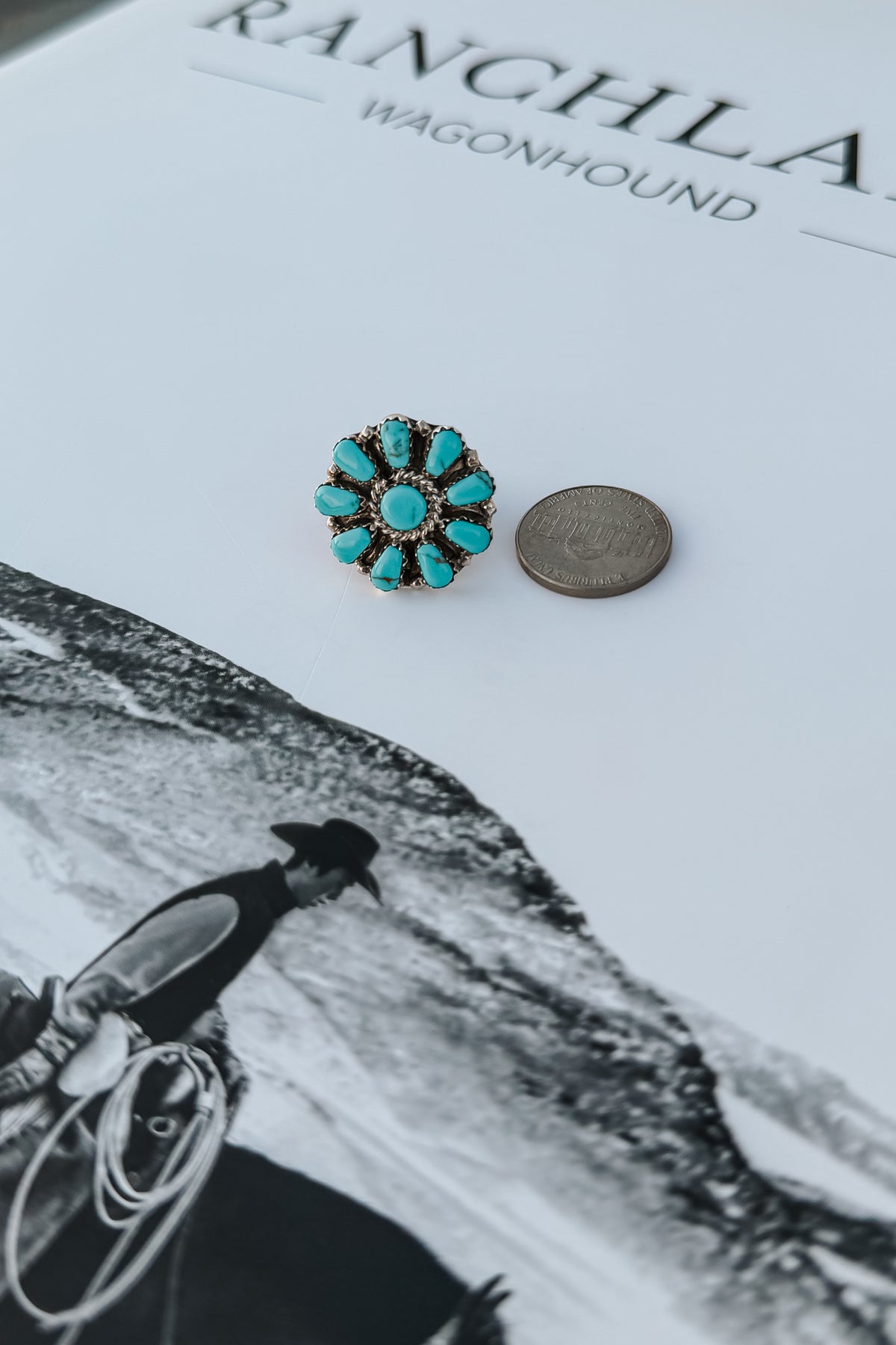 A single turquoise flower-shaped stud earring with a gold-toned setting and small clear stones, placed next to a coin on a white printed surface with black text and a black-and-white western scene.