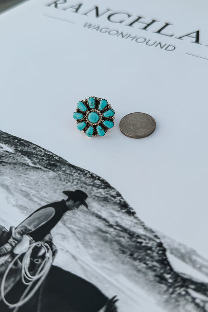 A single turquoise flower-shaped stud earring with a gold-toned setting and small clear stones, placed next to a coin on a white printed surface with black text and a black-and-white western scene.