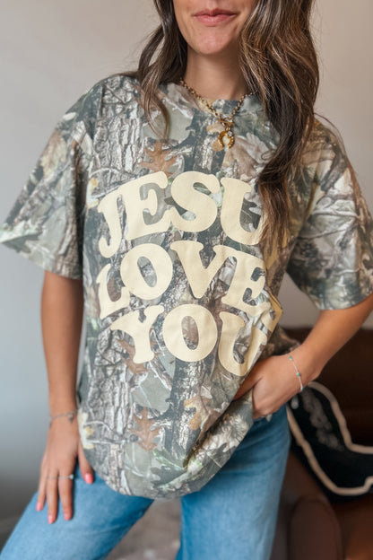 Model wearing oversized camouflage graphic t-shirt featuring “Jesus Loves You” text
