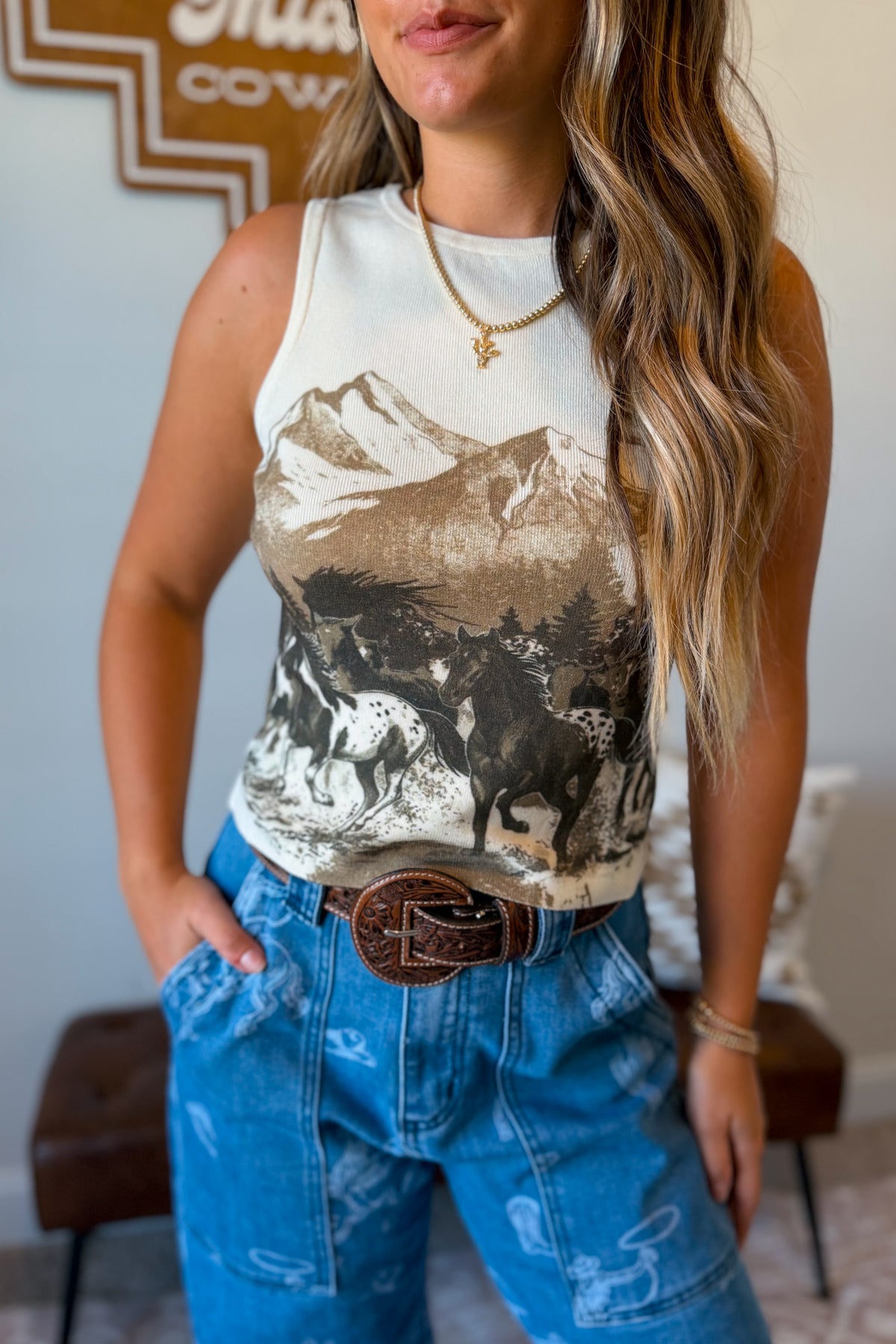 A woman wearing a sleeveless white tank top with a brown and black western mountain and horse graphic, paired with blue jeans and a brown belt.