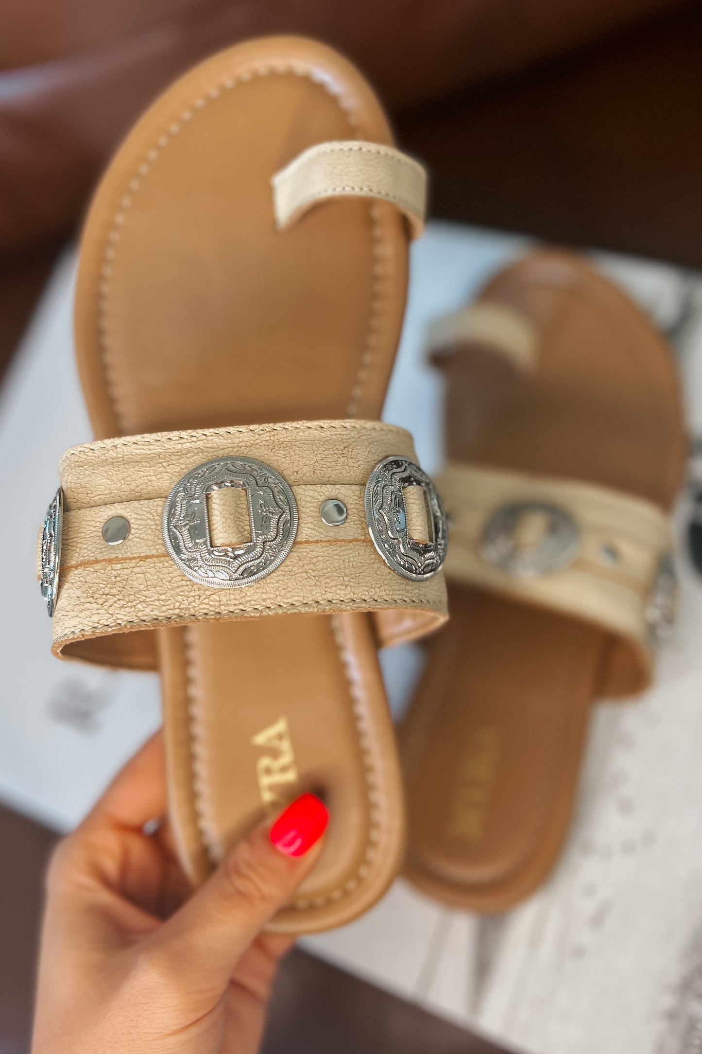Cream western slide sandals with tan cushioned footbed and toe loop. Wide textured strap features silver engraved concho accents and metal studs. Brand name printed on footbed.