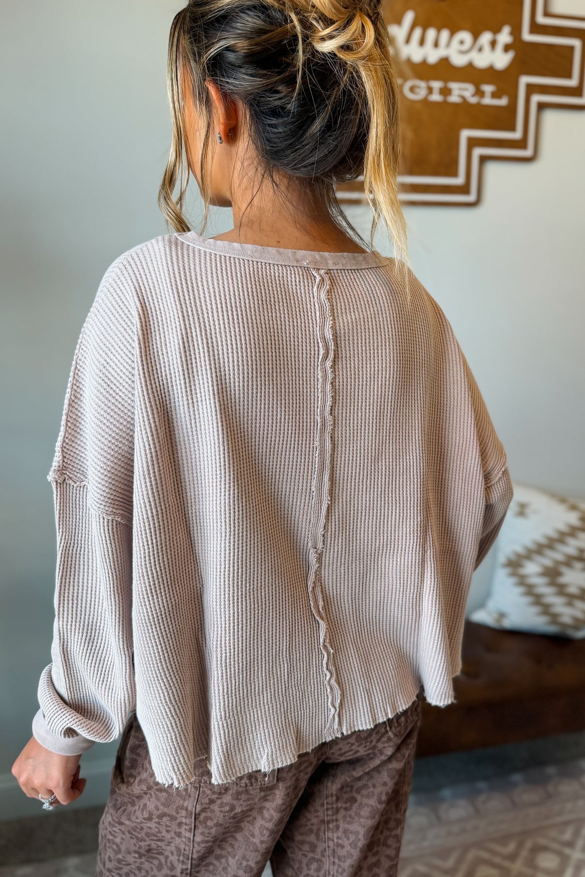 A woman shown from behind wearing a light beige ribbed knit long-sleeve top with a raw, frayed back seam detail, paired with brown patterned pants in an indoor setting.
