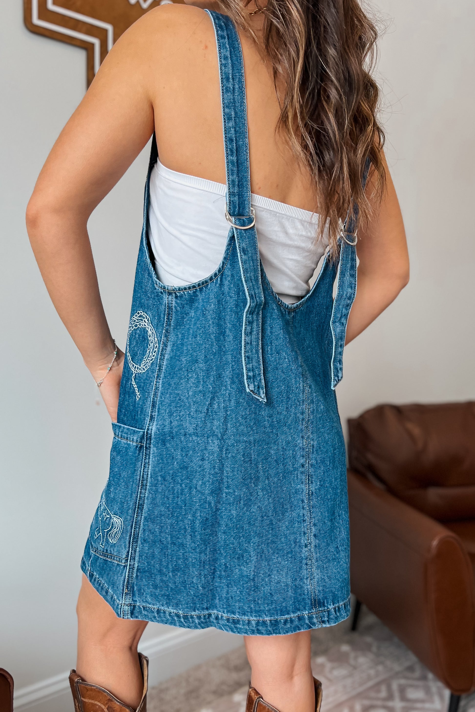 Back view of a blue denim overall dress showing adjustable straps and a straight hemline.