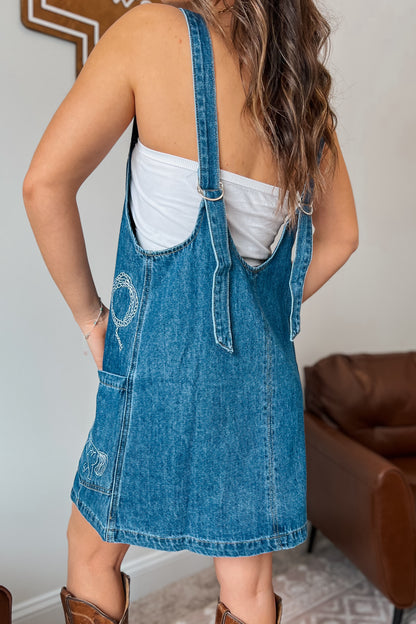 Back view of a blue denim overall dress showing adjustable straps and a straight hemline.