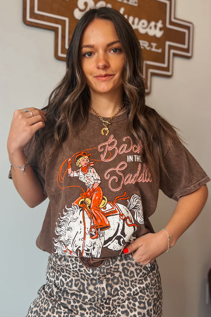 Brown vintage-wash graphic tee featuring a cowgirl riding a horse with “Back in the Saddle” text, styled with leopard print bottoms.