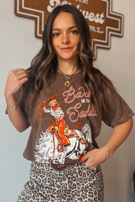 Brown vintage-wash graphic tee featuring a cowgirl riding a horse with “Back in the Saddle” text, styled with leopard print bottoms.