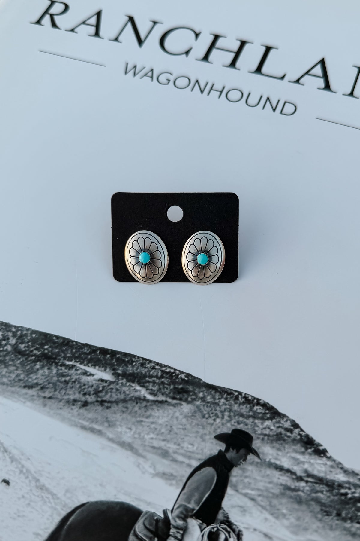 Silver-toned oval stud earrings with turquoise centers and floral detailing, displayed on a black card against a white background