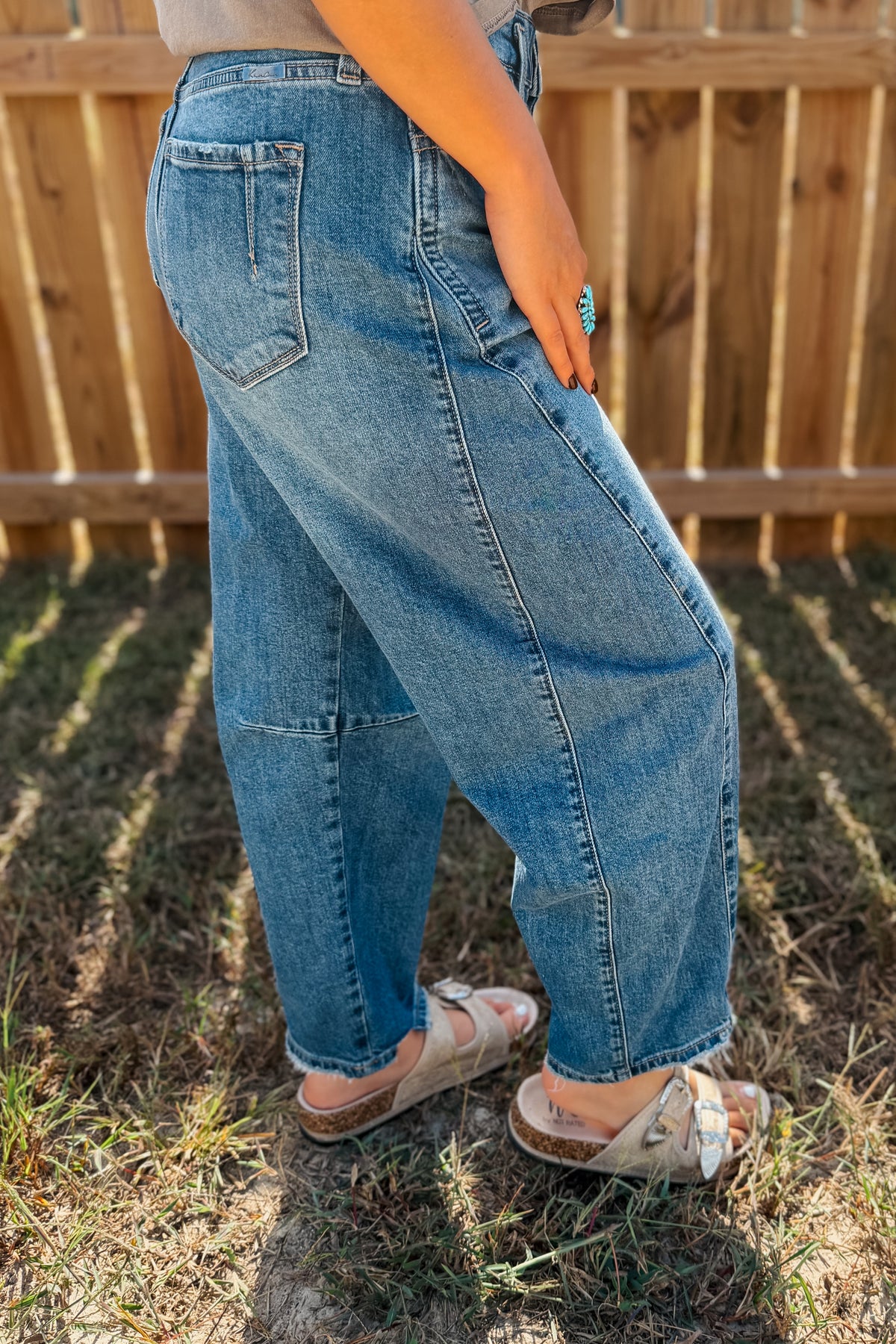 A person wearing loose, wide-leg light blue denim jeans and white slide sandals, standing outdoors on grass in front of a wooden fence.