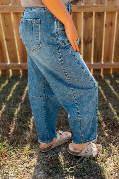 A person wearing loose, wide-leg light blue denim jeans and white slide sandals, standing outdoors on grass in front of a wooden fence.