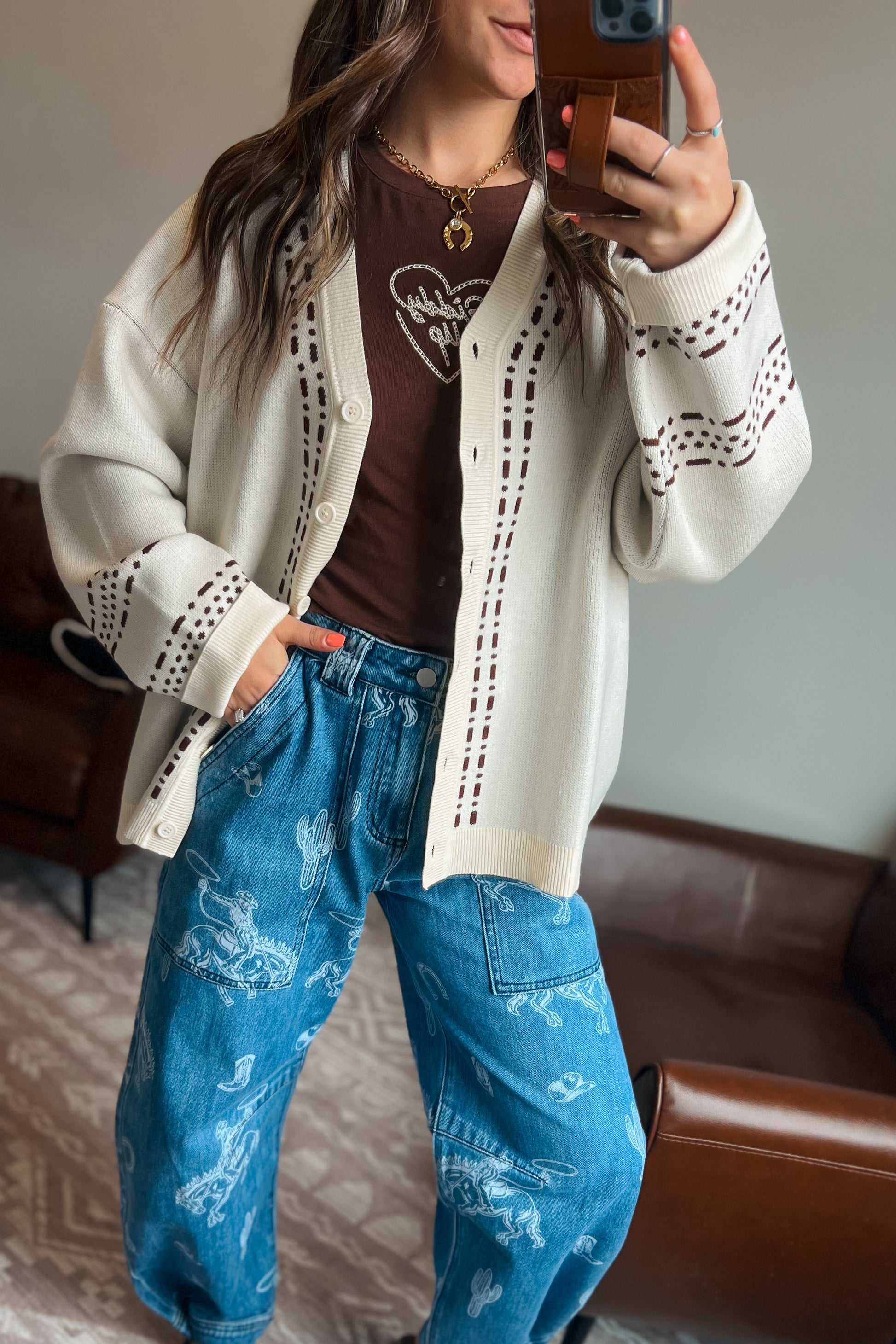 Model wearing a cream knit cardigan with brown western-inspired stitch detailing over a brown graphic tee and blue western print jeans.