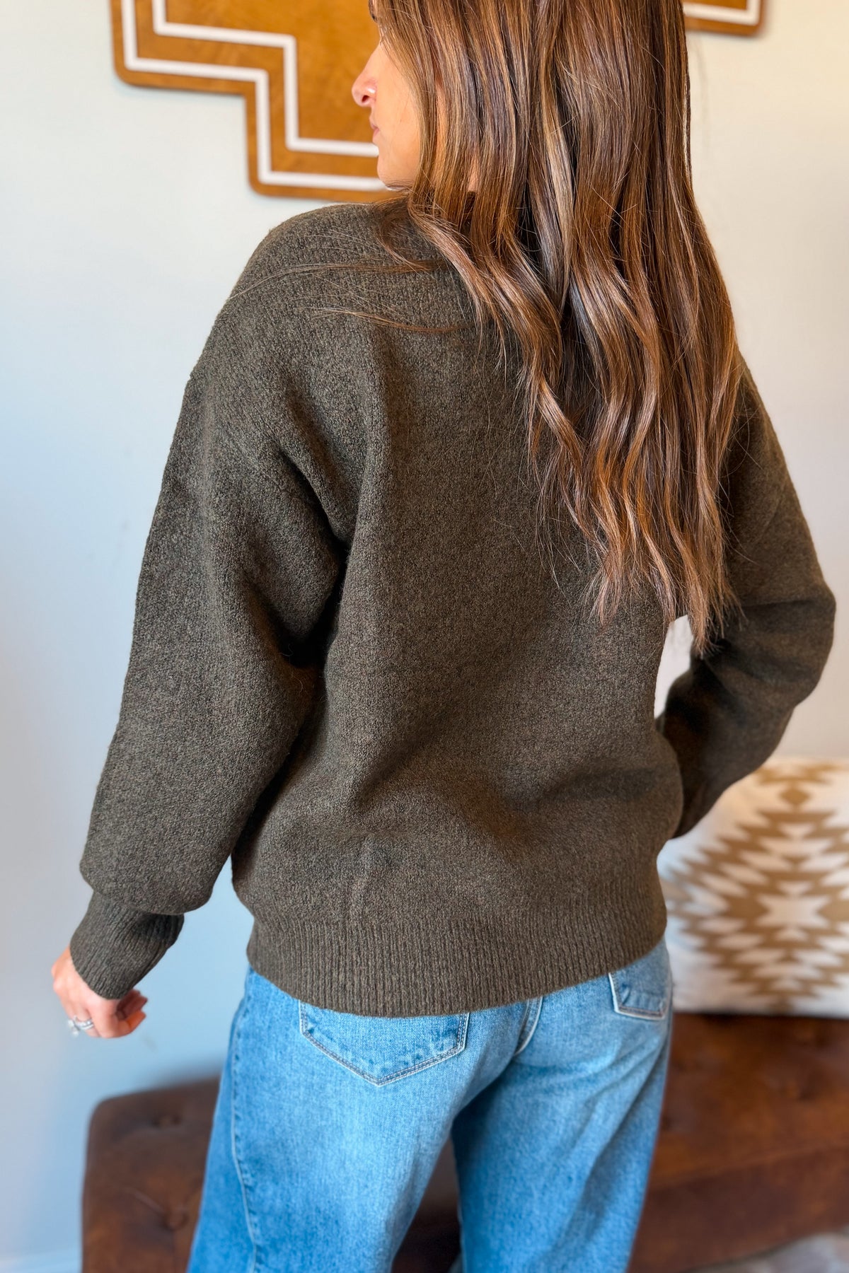 A woman wearing a dark olive brown knit sweater and blue jeans, shown from the back in an indoor setting.
