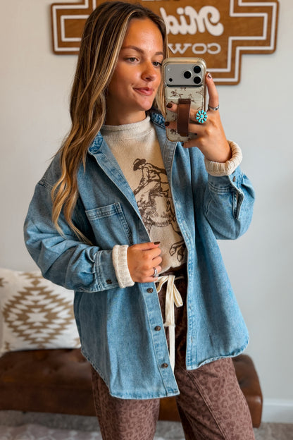 A woman takes a mirror selfie wearing a light blue denim button-up shirt over a beige graphic t-shirt, paired with brown patterned pants. The background shows a home interior with a brown leather sofa and a wooden sign.